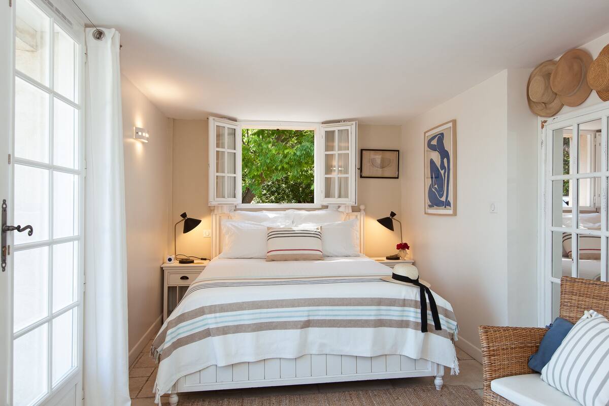 Ein Schlafzimmer mit einem Bett, zwei Nachttischen, einem fenster mit Vorhängen, einem Korbstuhl und einem an der Wand befestigten Hutfächer.