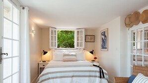 A bedroom with a bed, two bedside tables, a window with curtains, a wicker chair, and a wall-mounted hat rack. - Villa Le Port d'Attache (Cannes)