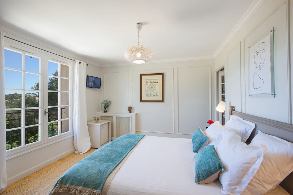 Ein Schlafzimmer mit einem großen Bett, einem fensterbelaubten Fenster, einem gerahmten Bild an der Wand und einer Deckenleuchte.