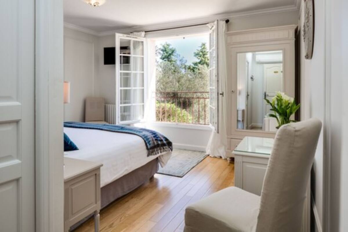 Ein Schlafzimmer mit Bett, Stuhl, Fenster mit Blick auf Grünflächen und einer Tür zu einem anderen Raum.