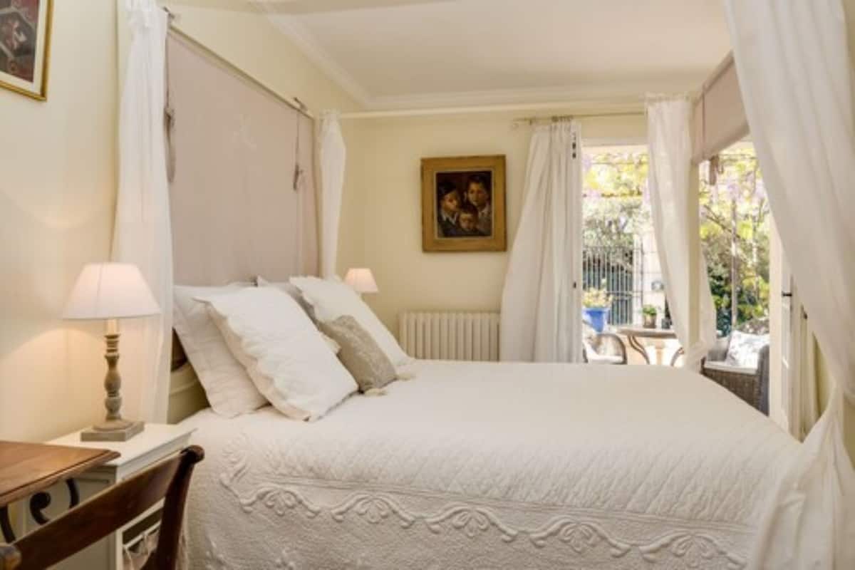 Ein Schlafzimmer mit Bett, zwei Nachttischlampen, einer Heizung, einem gerahmten Bild an der Wand und Blick ins Freie durch ein Fenster mit Vorhängen.