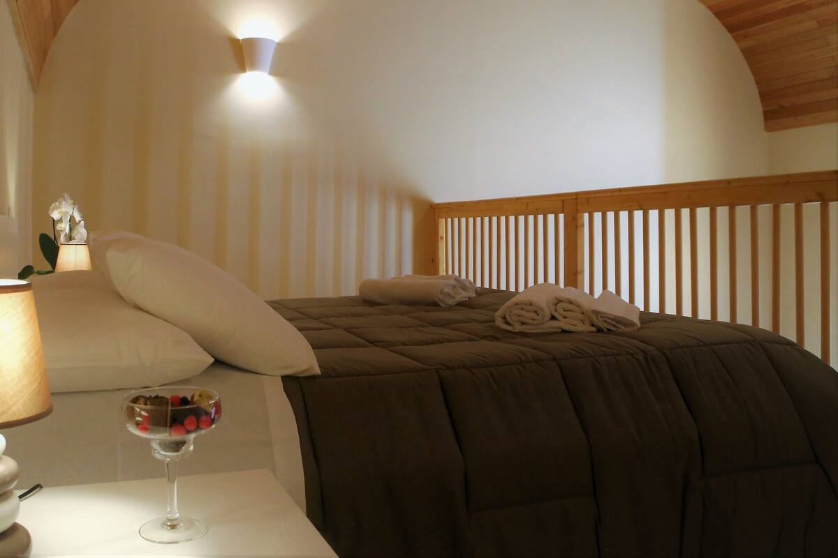 A bedroom with a wooden ceiling, a bed with a brown duvet, a bedside table with a glass of fruit, and a nightstand with a lamp.