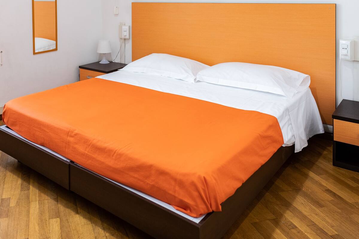 A bed with an orange bedspread and white pillows, a wooden headboard, a nightstand with a lamp, and a wooden floor.