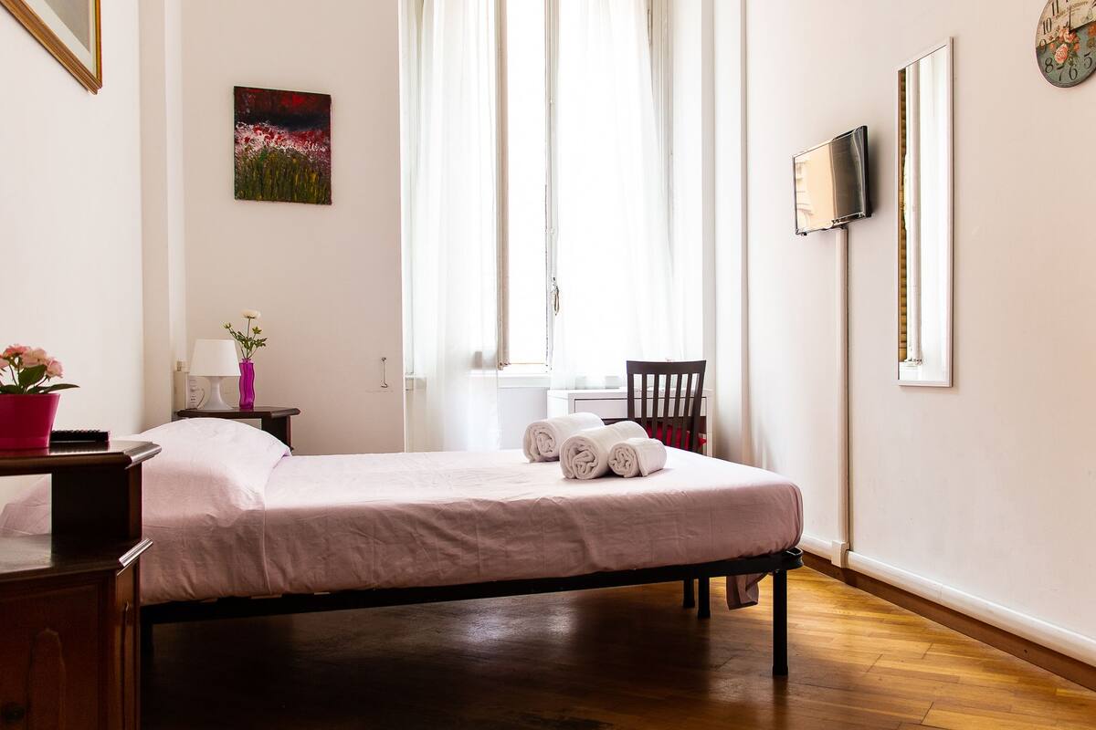 A neatly made bed with rolled towels, a bedside table with a lamp and a vase, a chair, and a painting on the wall.