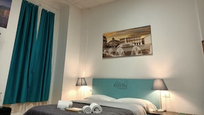 A hotel room with a bed, two bedside lamps, a painting of a cityscape, teal curtains, and a headboard with a message. - Hotel Youri Il Magnifico (Genoa)