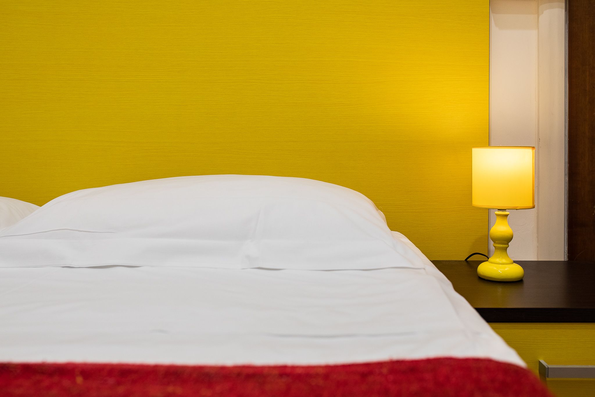 Un letto con biancheria bianca e una coperta rossa, una lampada gialla su un comodino e una parete gialla.
