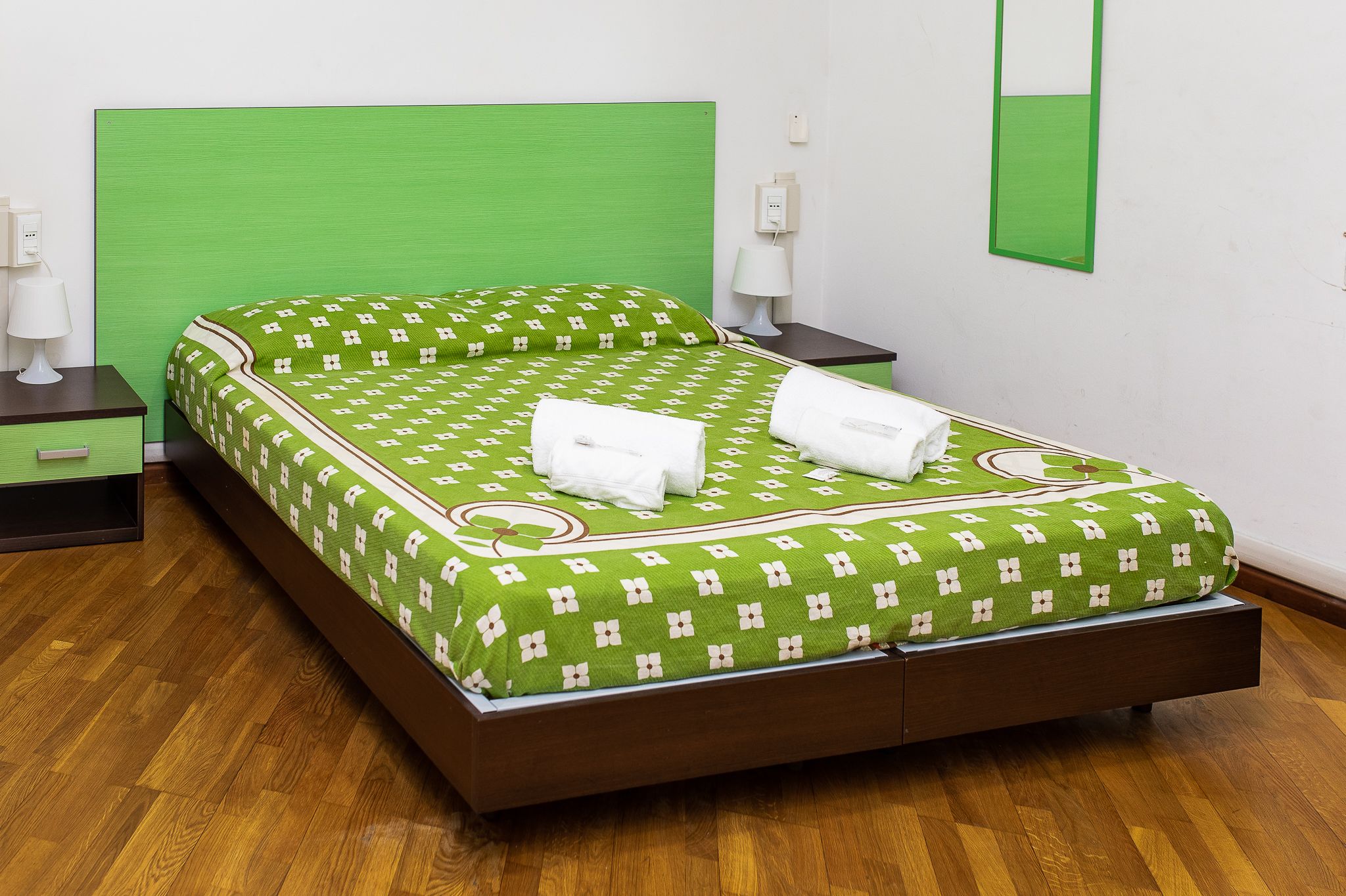 Ein ordentlich bezogenes Bett mit grünem Kopfteil und gemusterten Bettwäsche, zwei Nachttische mit Lampen und ein Holzboden.