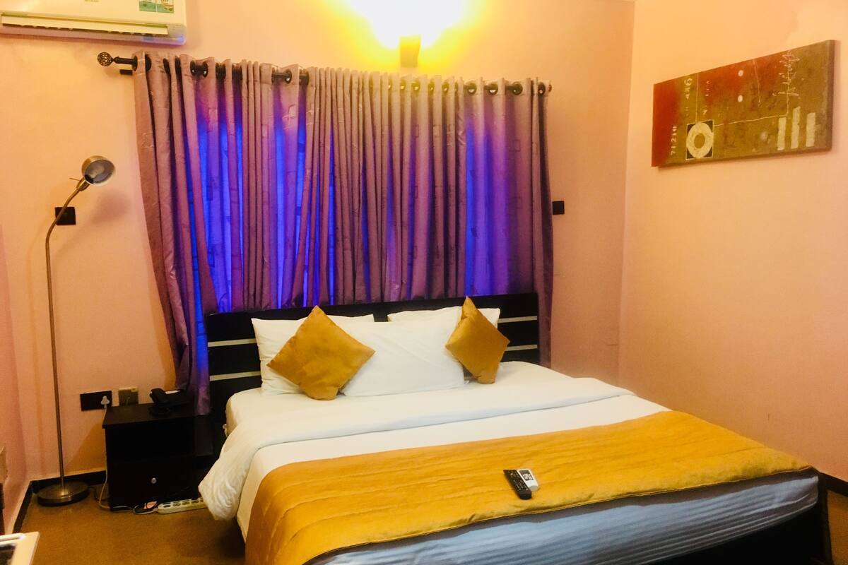 A hotel room with a bed, a bedside table, a lamp, and a wall-mounted air conditioner.