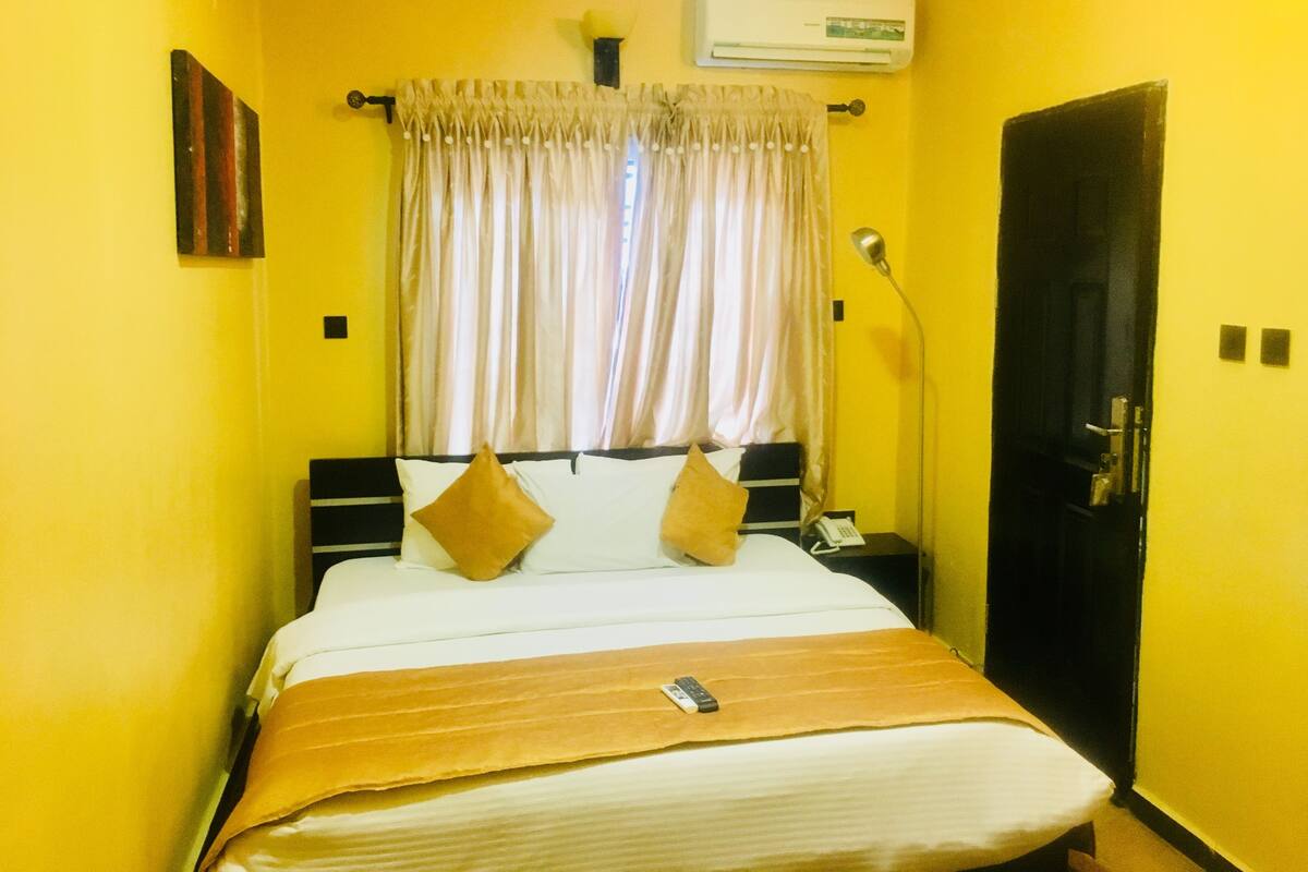 A hotel room with a bed, curtains, an air conditioning unit, a bedside lamp, and a door.