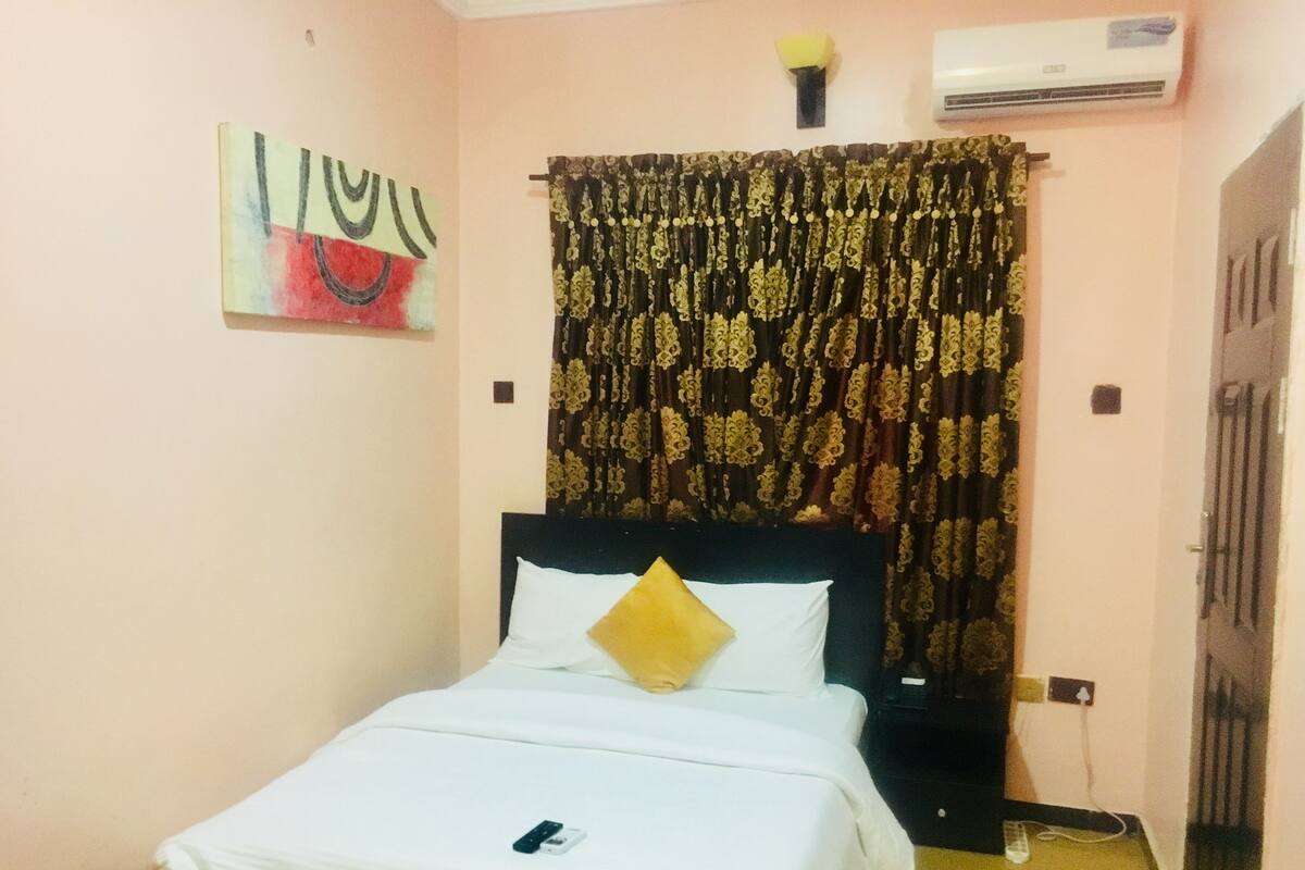 A neatly made bed with white bedding and a single yellow pillow, a wall-mounted air conditioner, and a decorative curtain with a floral pattern.