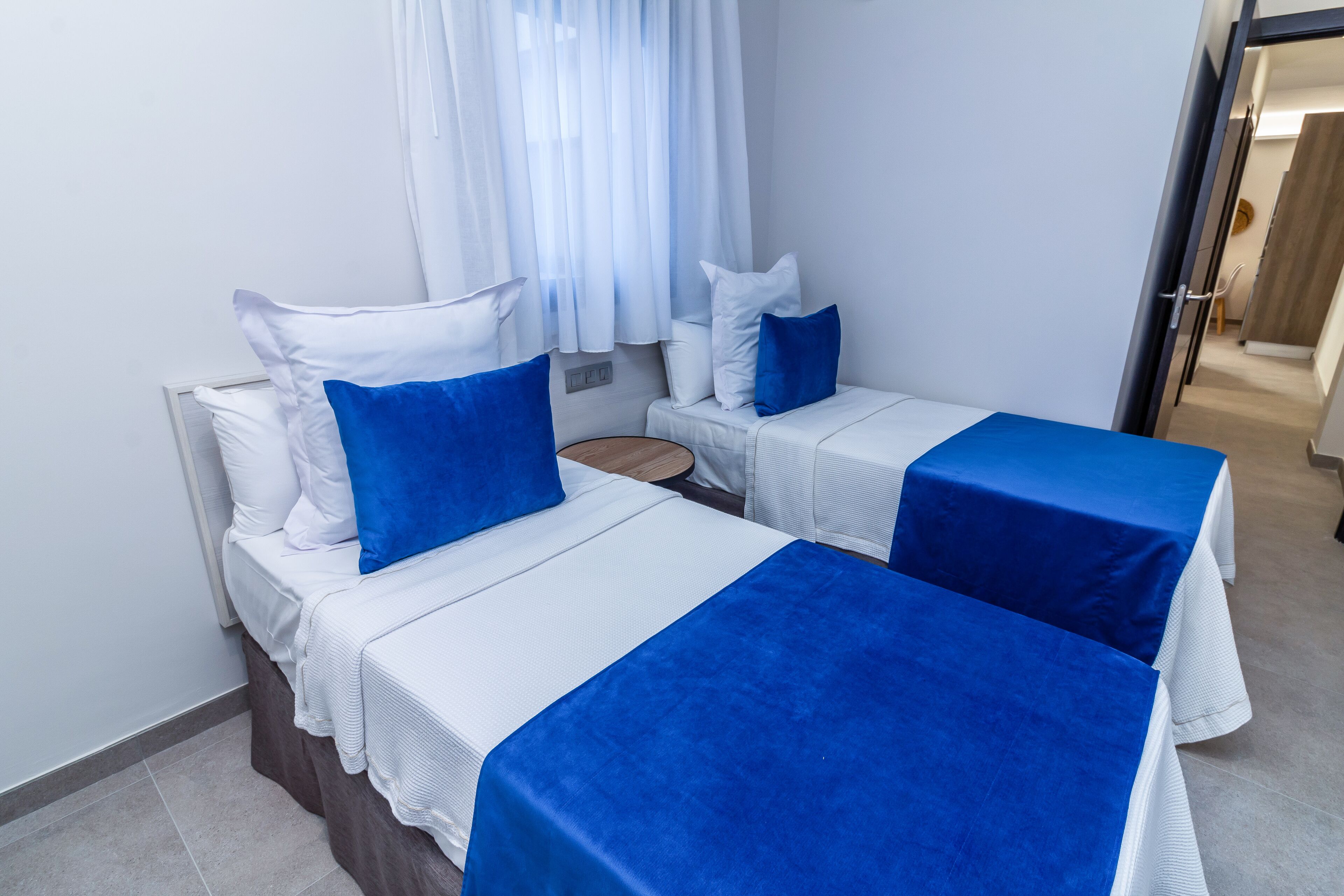 A hotel room with two single beds, each with a blue bedspread and white pillows. There is a small round table between the beds, and the room has white walls and a window with white curtains.