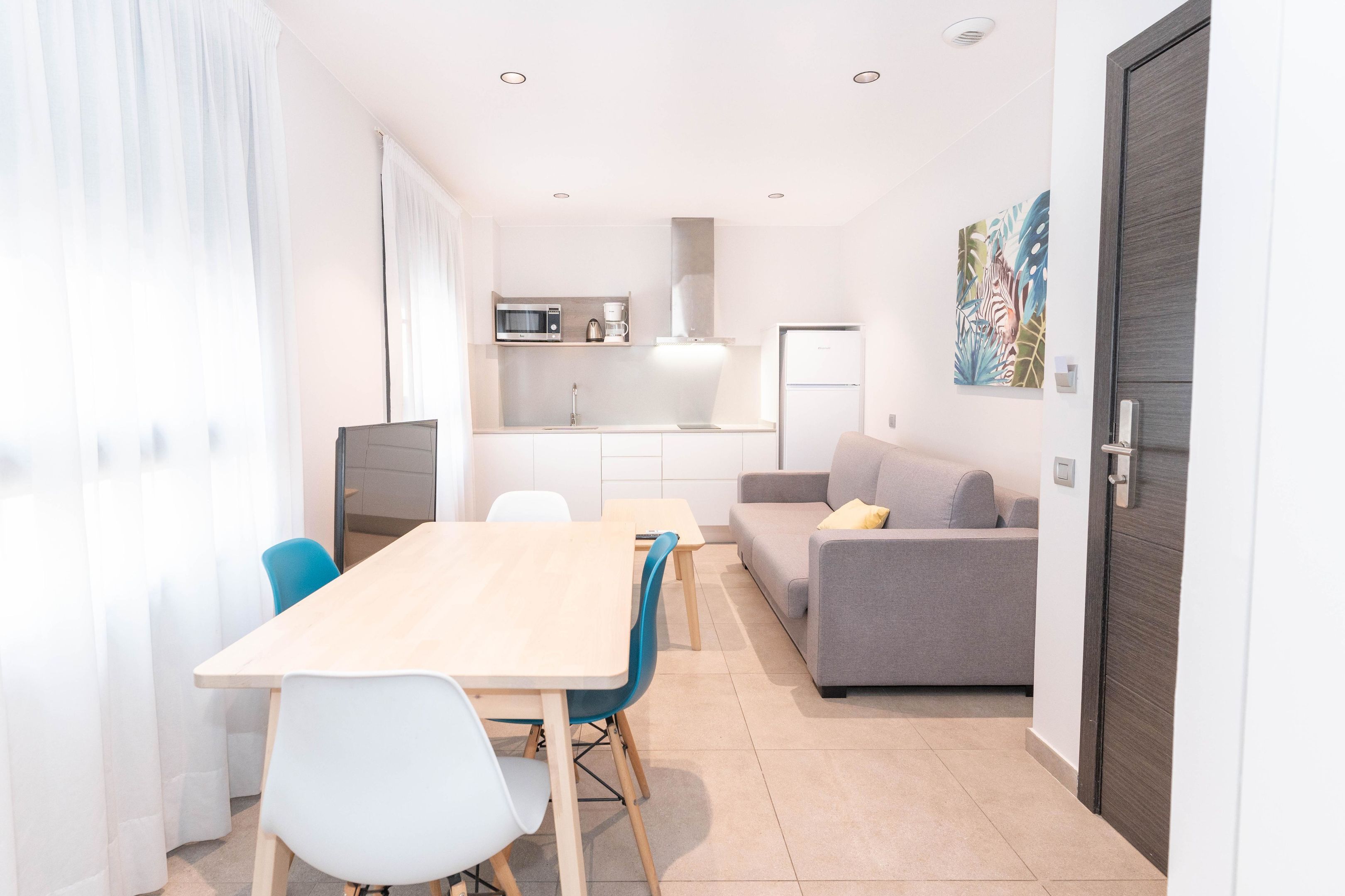 A modern kitchen with a microwave, coffee maker, and refrigerator. There is a dining area with a table and chairs, and a gray sofa with a yellow pillow.