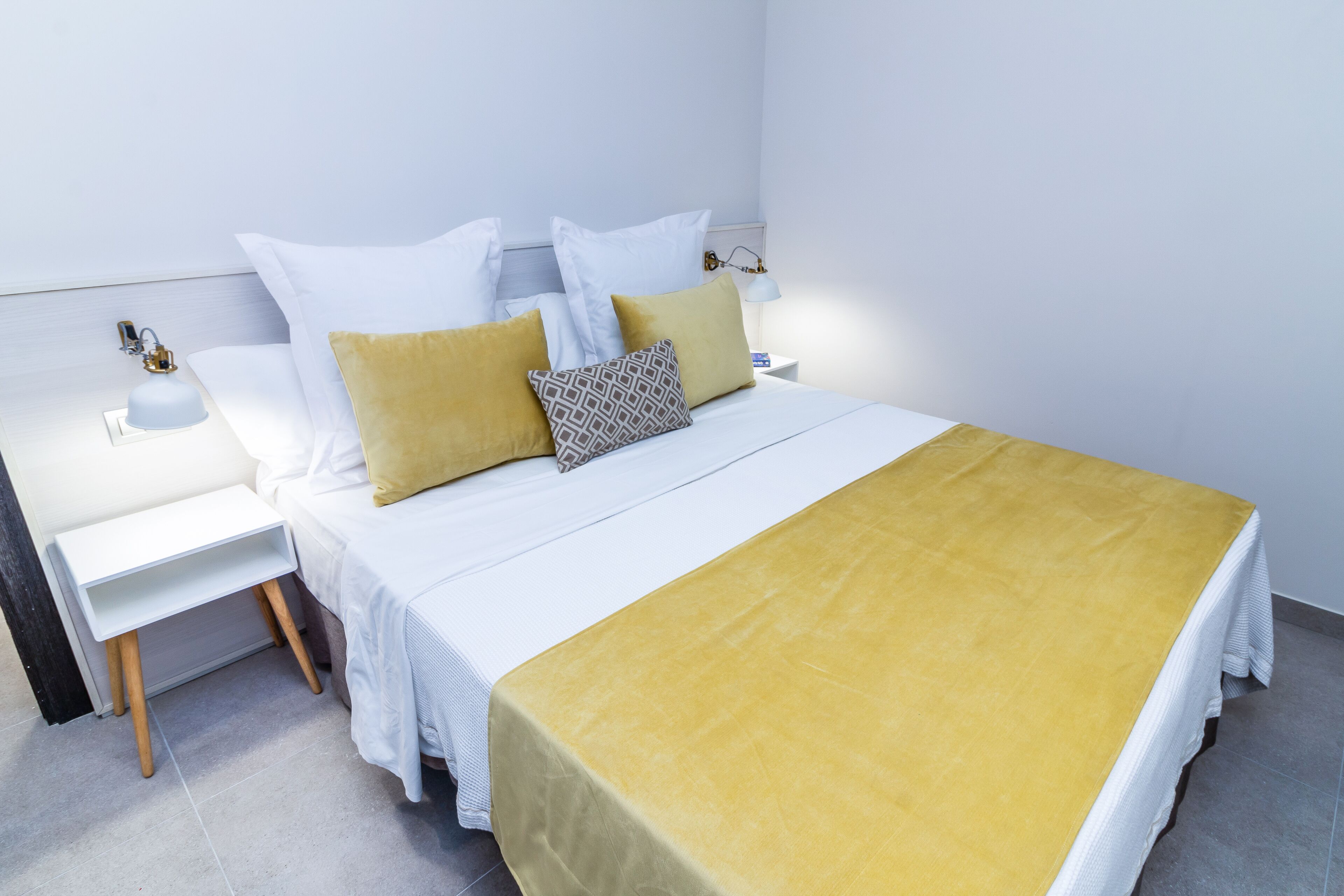 Een net opgemaakt bed met witte beddengoed en een mosterdgele plaid, een nachtkastje met een lamp, en een wandlamp.