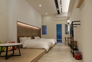 A modern hotel room with a large bed, a small table, and a desk. There is a window with a view of trees and a balcony.