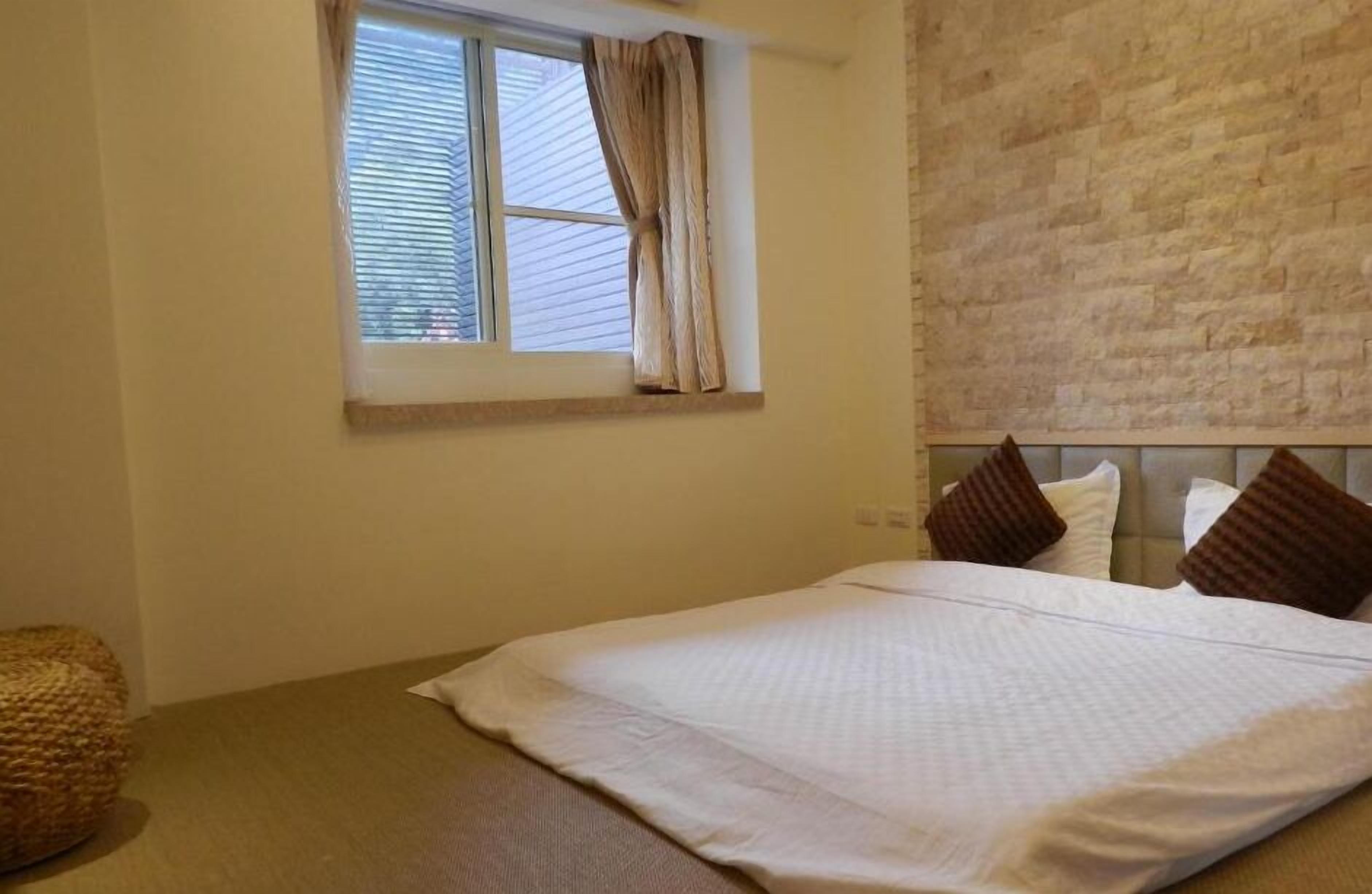 Een slaapkamer met een bed, een raam met gordijnen, een stenen muur en een geweven stoel.