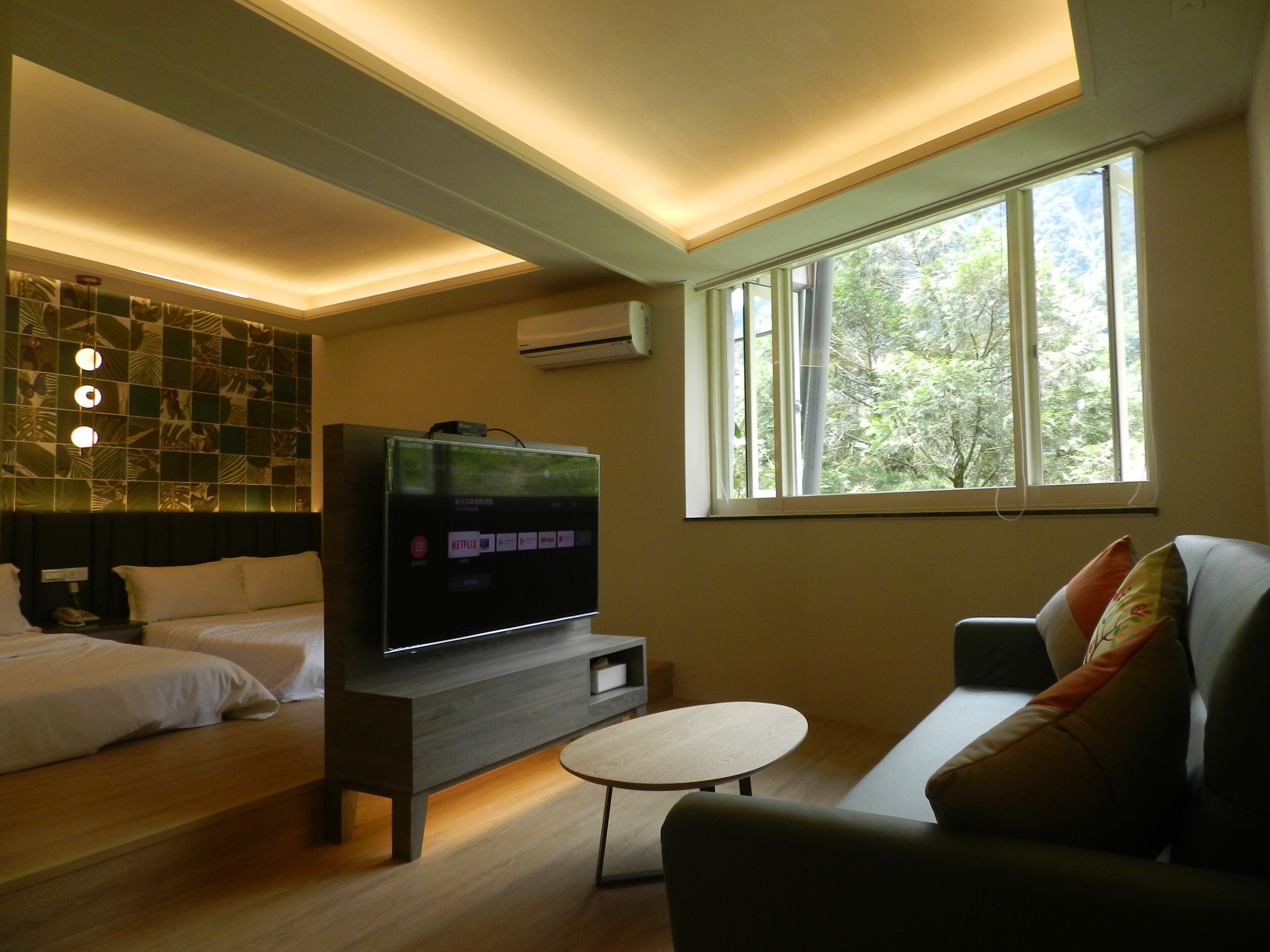 A modern hotel room with a large flat-screen TV, a sofa, a small round table, and a bed with two pillows.