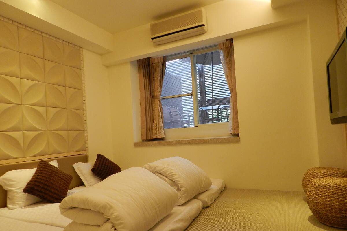 A hotel room with a bed, pillows, a window with curtains, an air conditioning unit, and a woven chair.
