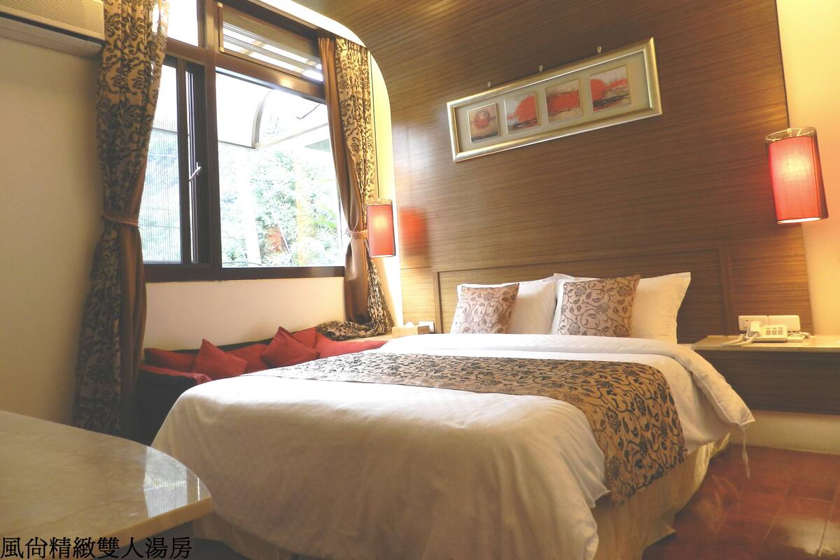 A hotel room with a bed, a bedside table, a lamp, a telephone, and a window with curtains.
