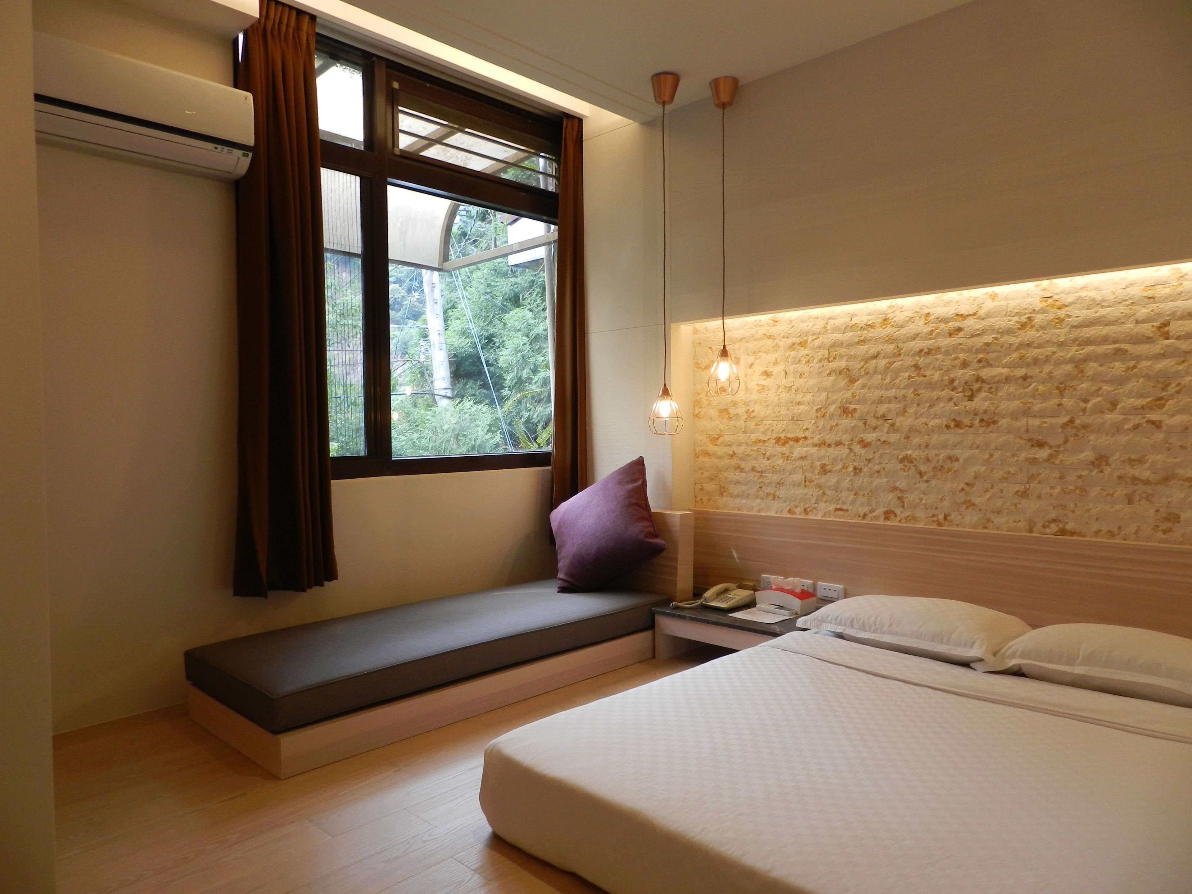 Un dormitorio con una cama, un banco, una ventana con cortinas, un aire acondicionado instalado en la pared y una pared con textura.