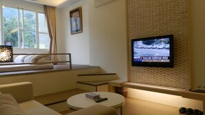 Ein Hotelzimmer mit Bett, Wandfernseher, Fenster mit Vorhängen und einer kleinen Sitzecke mit Tisch.