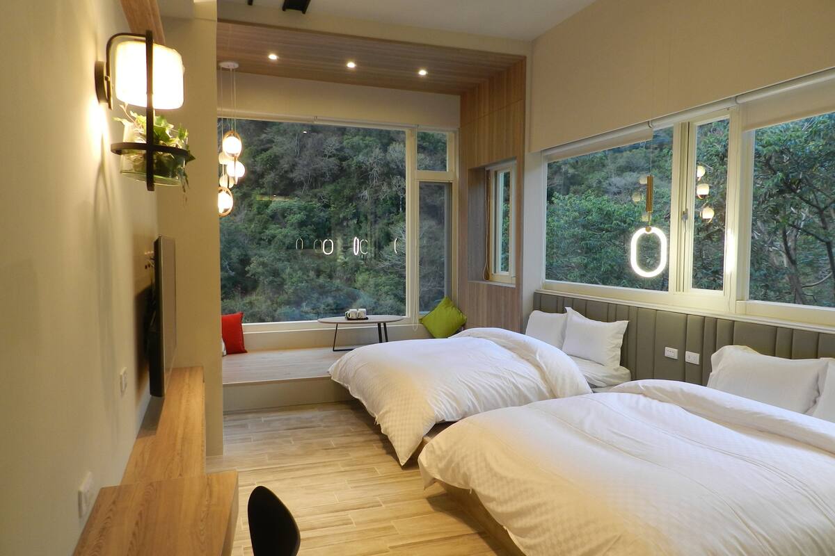 A modern bedroom with two beds, large windows, and a view of greenery.