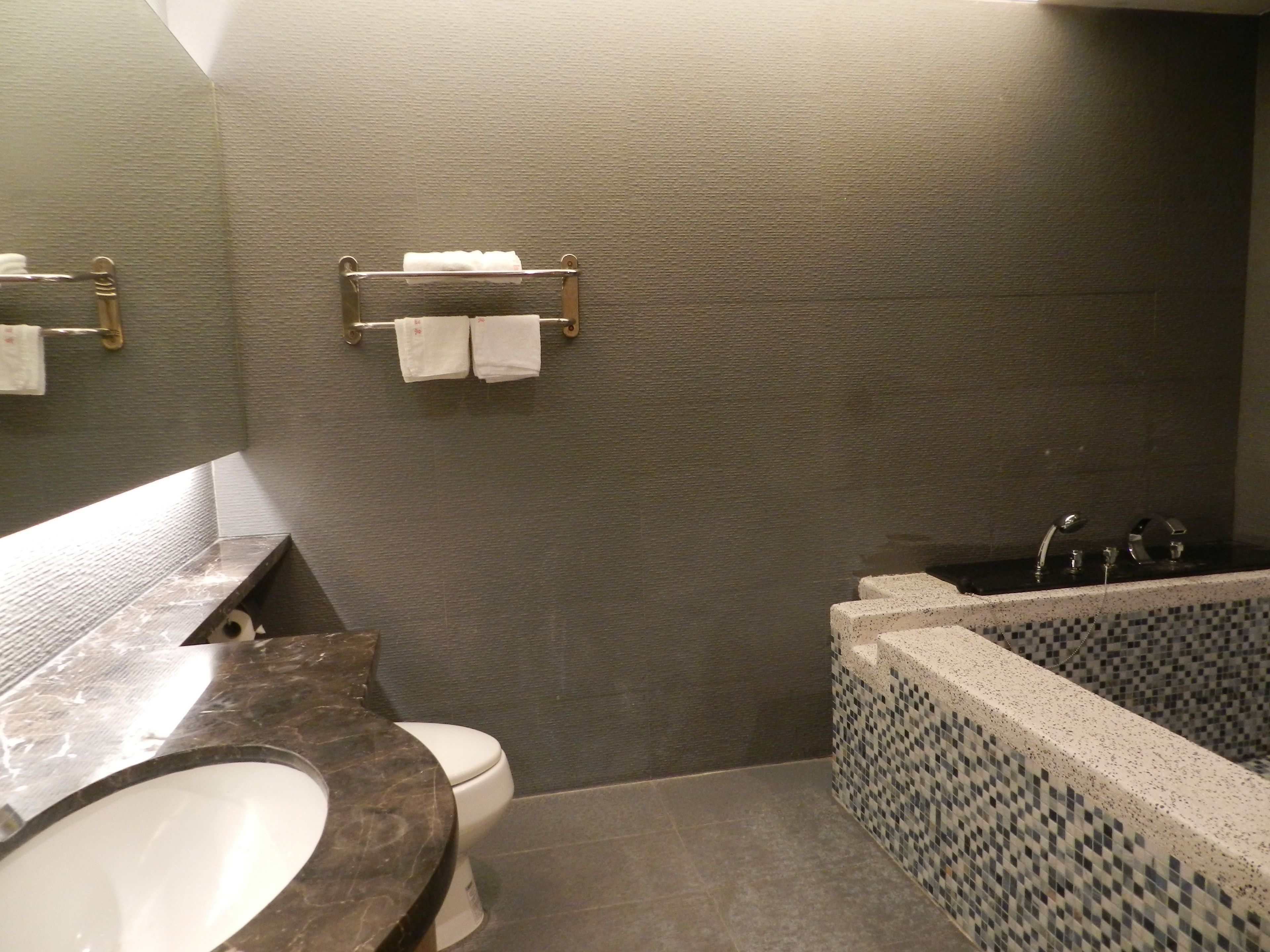 Un baño con lavamanos de mármol, inodoro, bañera con mosaicos y un perchero con dos toallas.