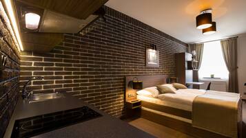A modern hotel room with a brick wall, a bed, a bedside table with a lamp, a built-in stove, and a window with curtains.