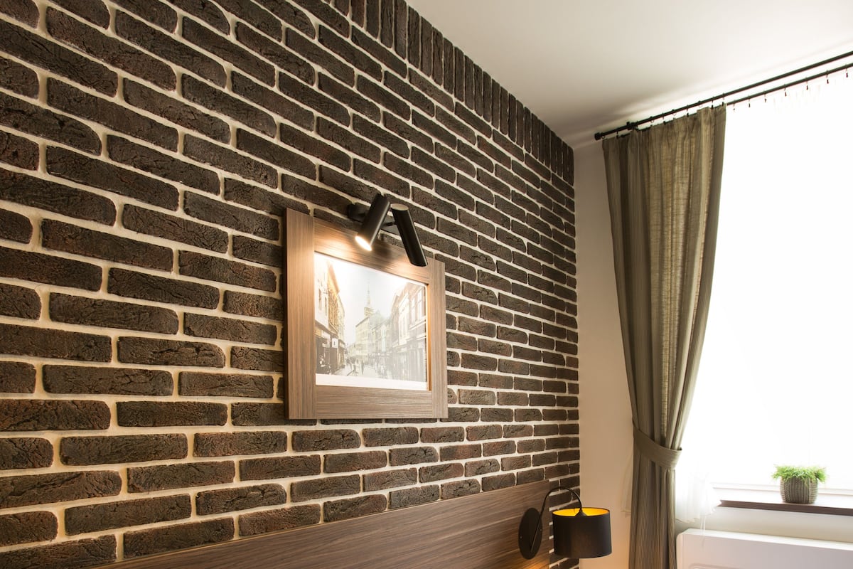 A hotel room with a brick wall, a framed picture, a bedside lamp, a bed with pillows, and a window with curtains.
