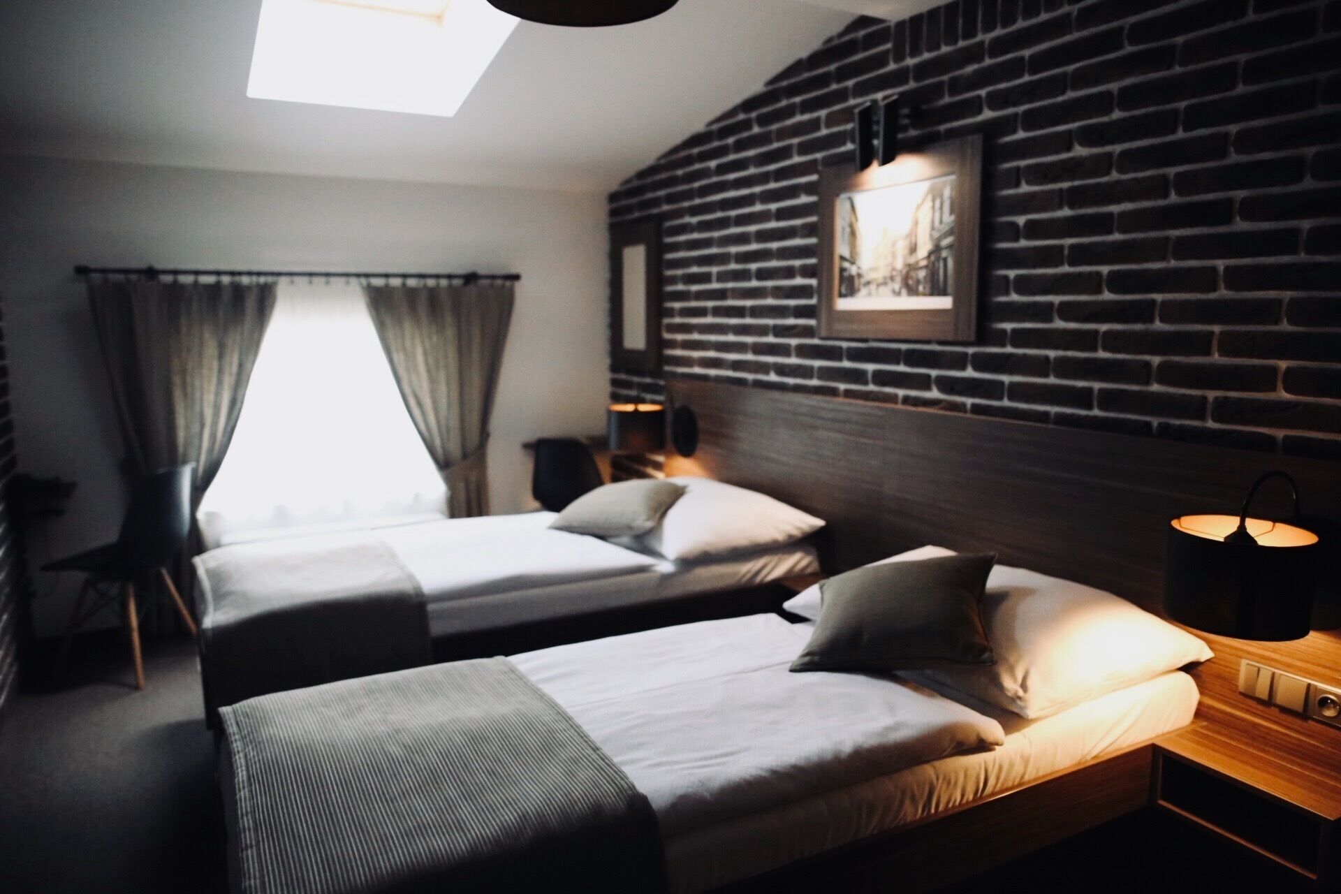A hotel room with two beds, a brick wall, a window with curtains, and a nightstand with a lamp.