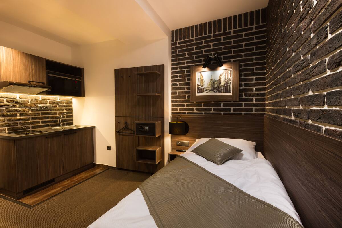A compact hotel room with a kitchenette, a bed with a headboard, a nightstand, and a wall-mounted picture.