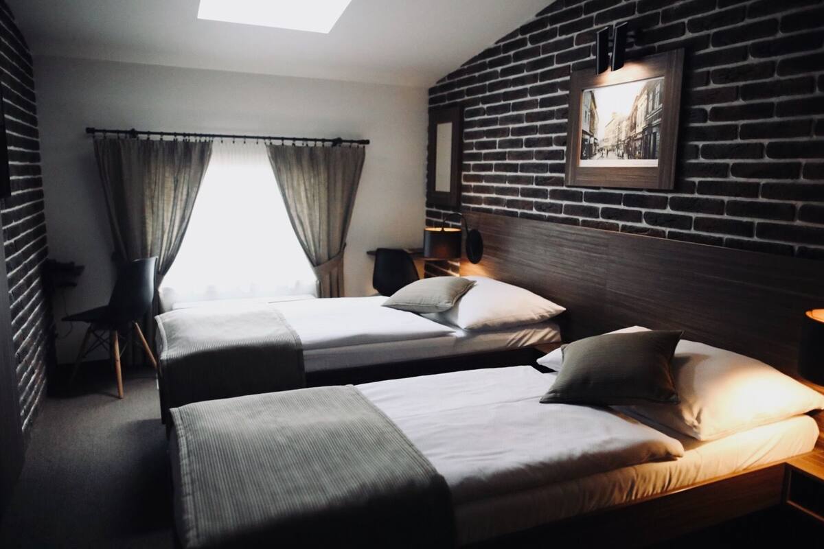 A hotel room with two beds, a brick wall, a large window with curtains, and a framed picture on the wall.