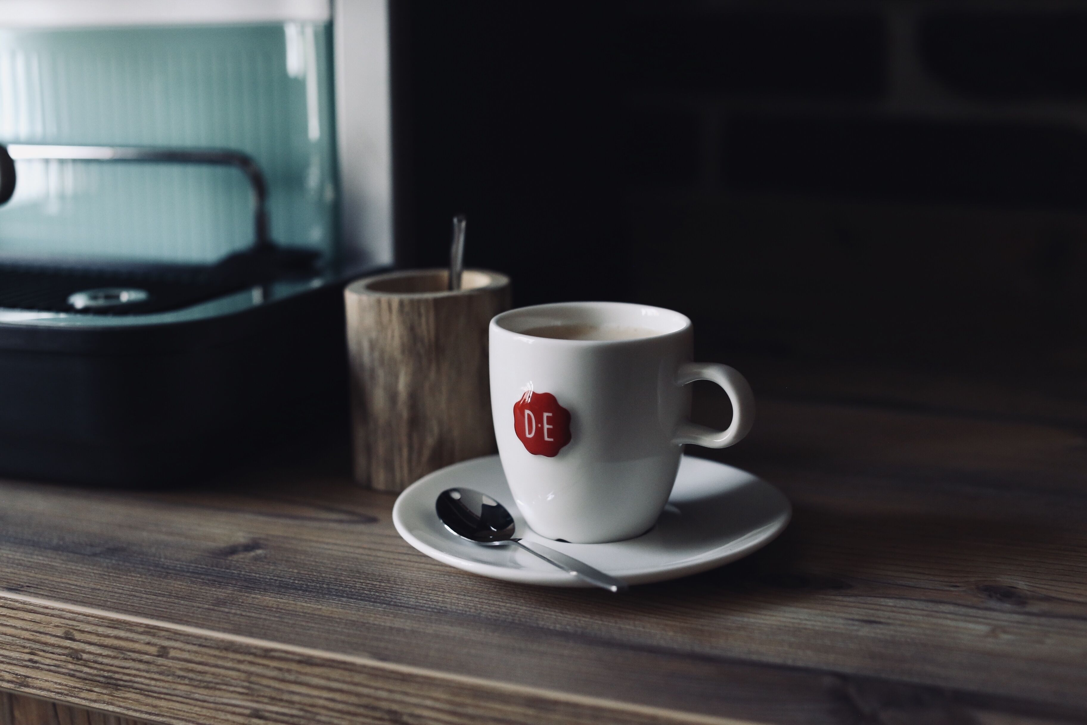A white cup with a red logo on a wooden table, accompanied by a spoon and a wooden cup.