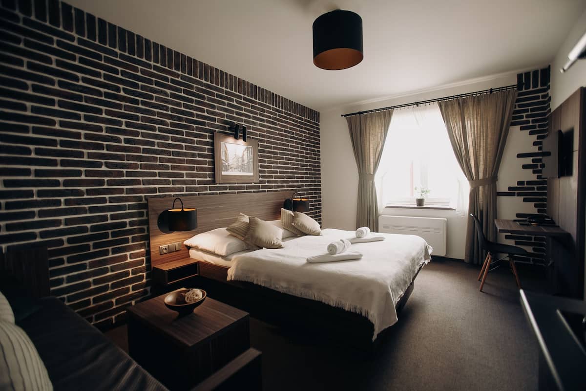 A modern hotel room with a brick-patterned wall, a neatly made bed, a bedside table with a lamp, and a view of the outdoors through a window.