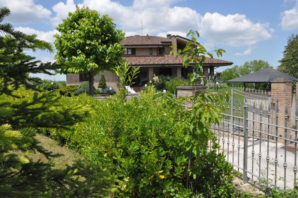 A two-story house with a tiled roof, surrounded by lush greenery and a metal fence.