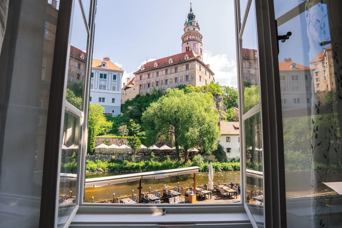 Blick aus einem Fenster auf ein Flussufer mit einem historischen Gebäude, Sitzgelegenheiten im Freien und einer Brücke.