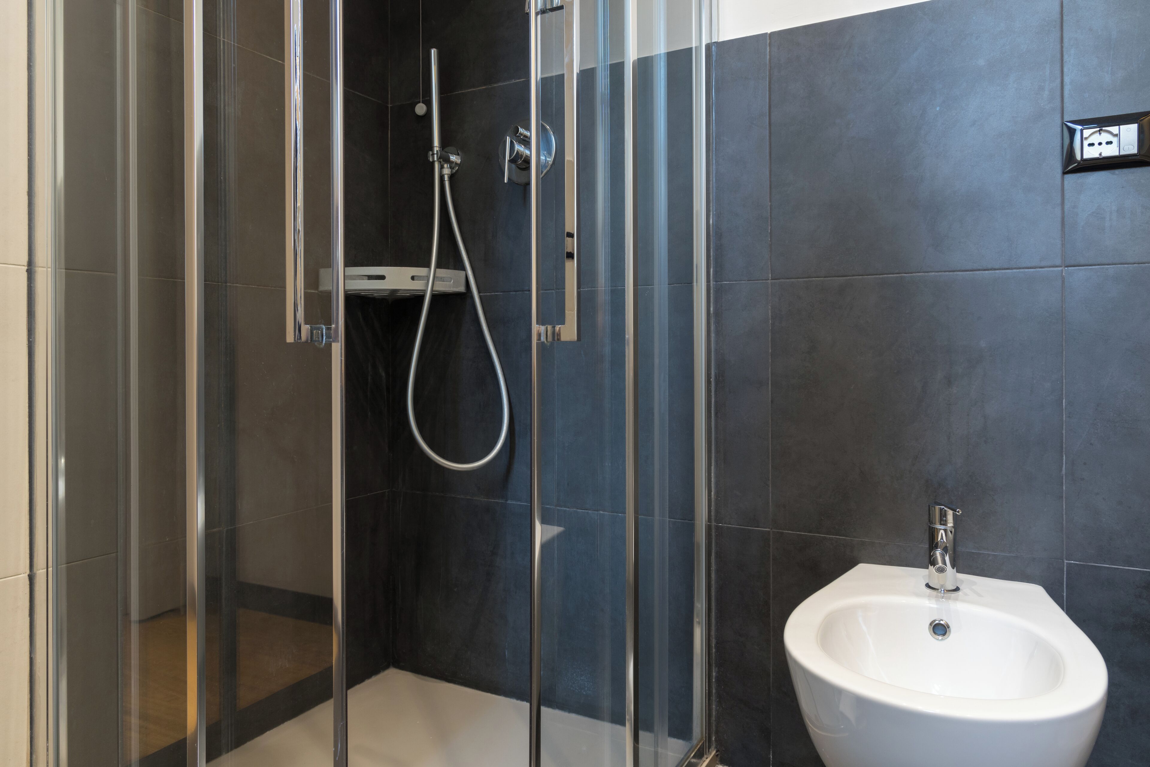 A modern bathroom with a glass-enclosed shower, a white sink, and dark tiled walls.