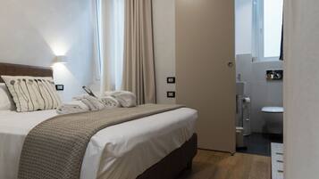 Ein Hotelzimmer mit Bett, Nachttisch, Lampe, einem Fenster mit Vorhängen und Blick ins Badezimmer.
