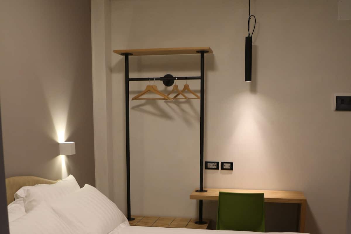 Una cama bien hecha con ropa blanca, un escritorio de madera con una silla, y un perchero en la pared.