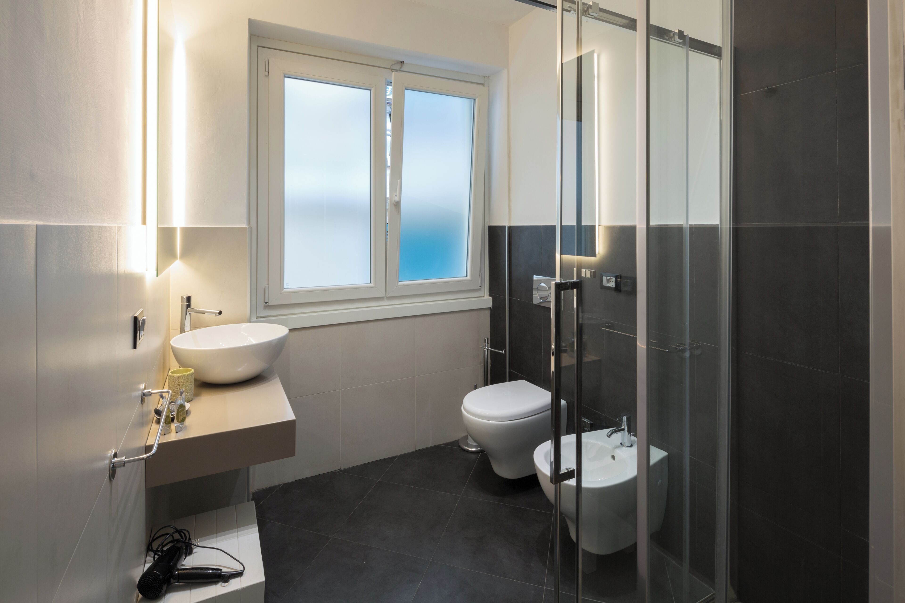 Un baño moderno con un ventanal grande, un lavabo, un inodoro y una cabina de ducha con mampara de vidrio.