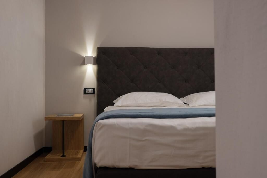 Een net opgemaakt bed met wit- en blauw beddengoed, een donker hoofdbord en een klein houten nachtkastje met een lamp.