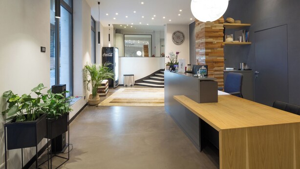 A modern reception area with a wooden reception desk, potted plants, and a staircase leading to an upper floor. - Hotel Doria (Genoa)