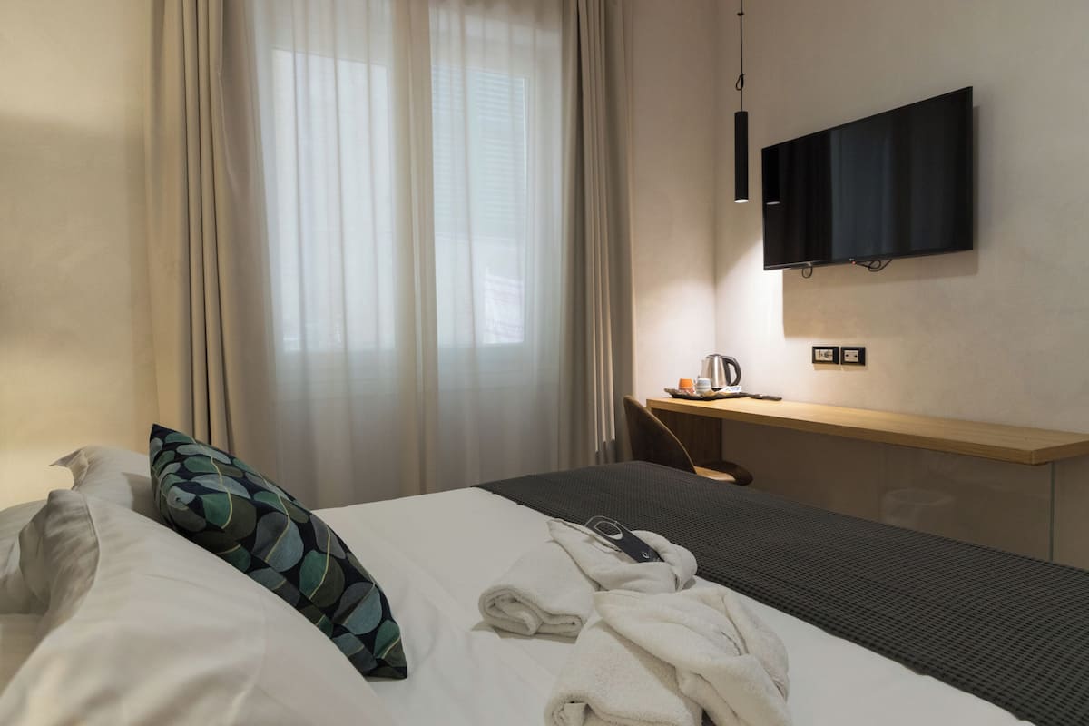 Una habitación de hotel con cama, escritorio, televisor y ventana con cortinas.