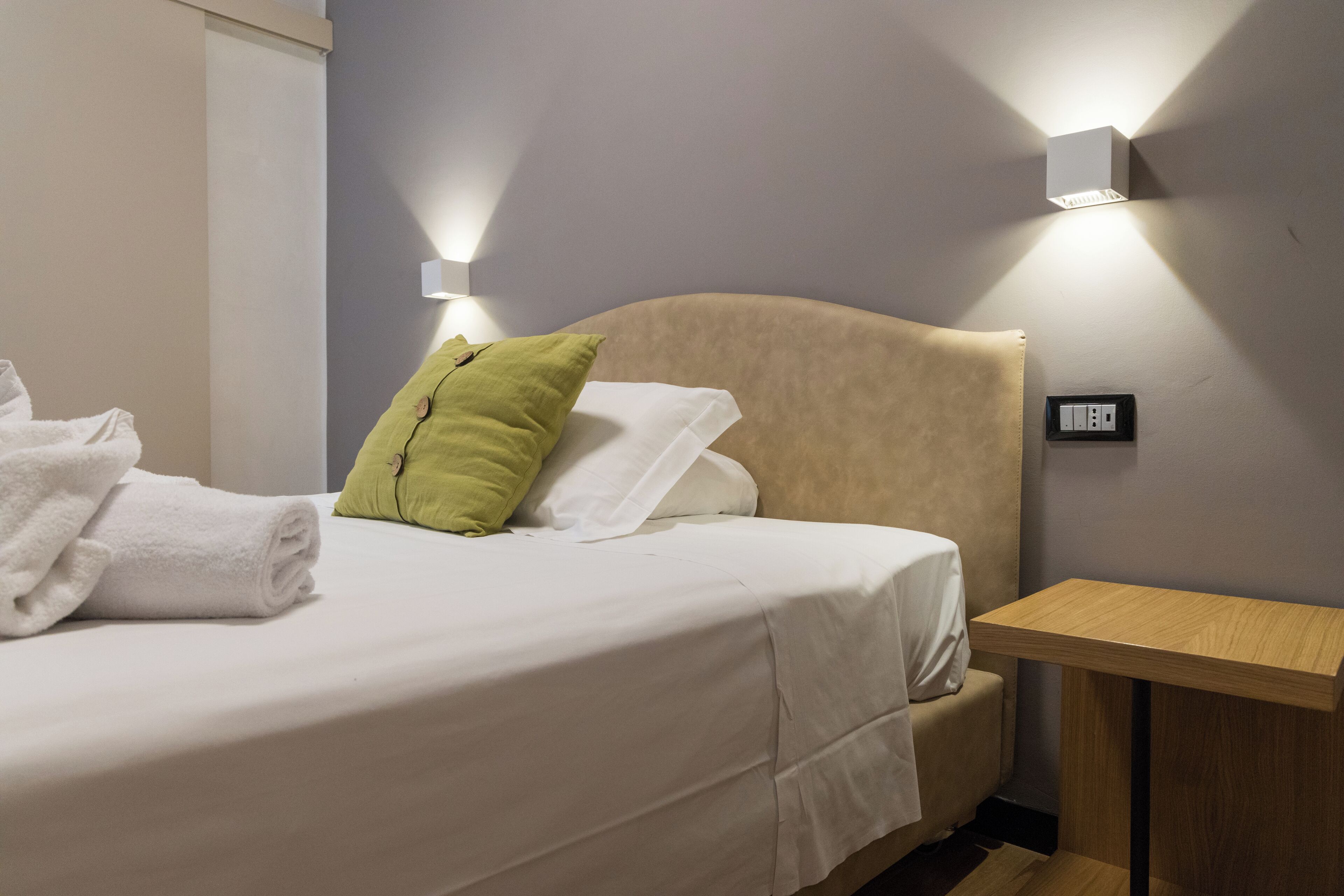 Un lit bien fait, avec des oreillers blancs et verts, une table de chevet en bois et un luminaire fixé au mur.