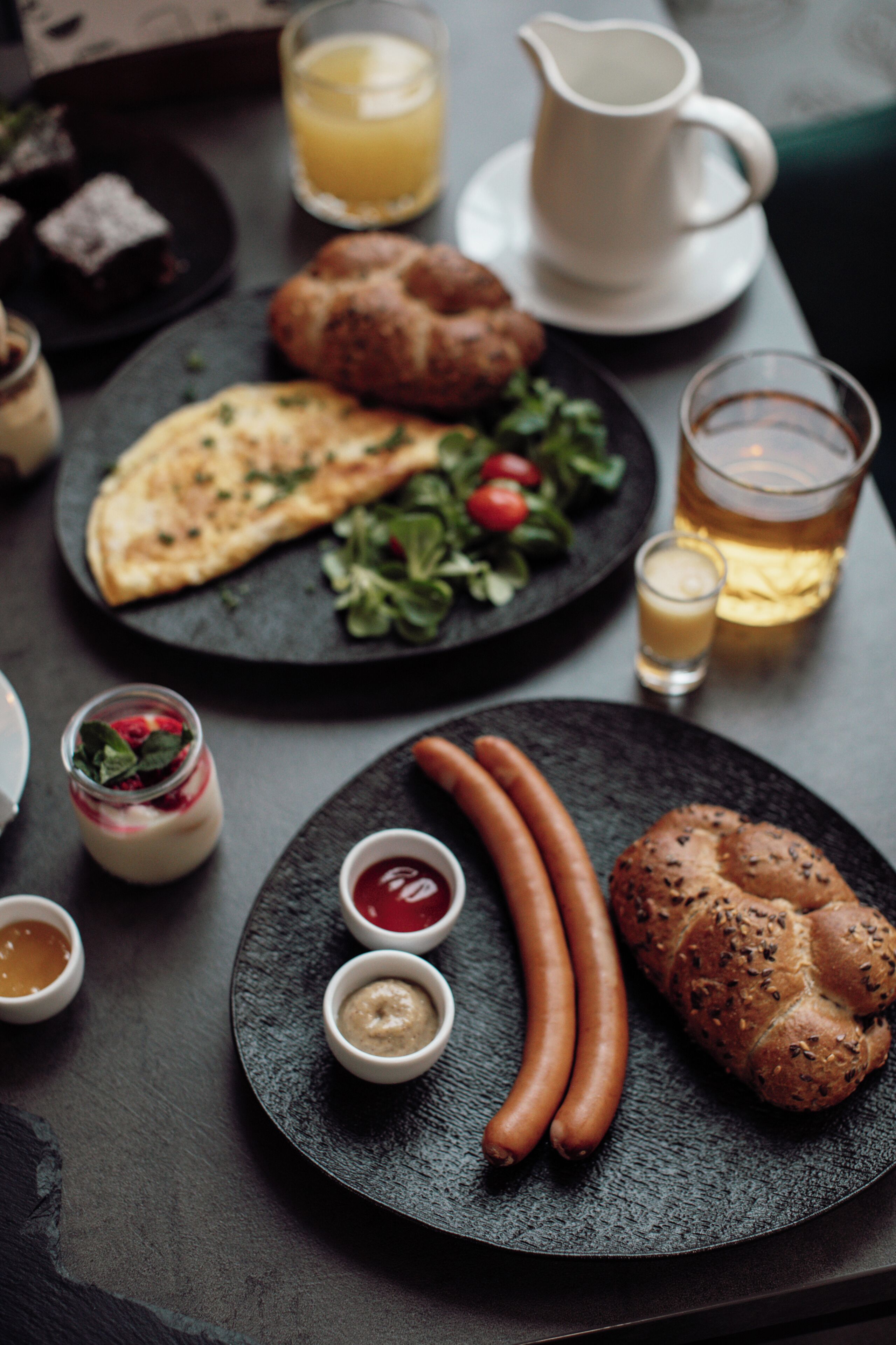 Una mesa de comedor con platos de comida, incluyendo salchichas, un croissant y una ensalada. También hay pequeños recipientes de condimentos y una jarra de agua.
