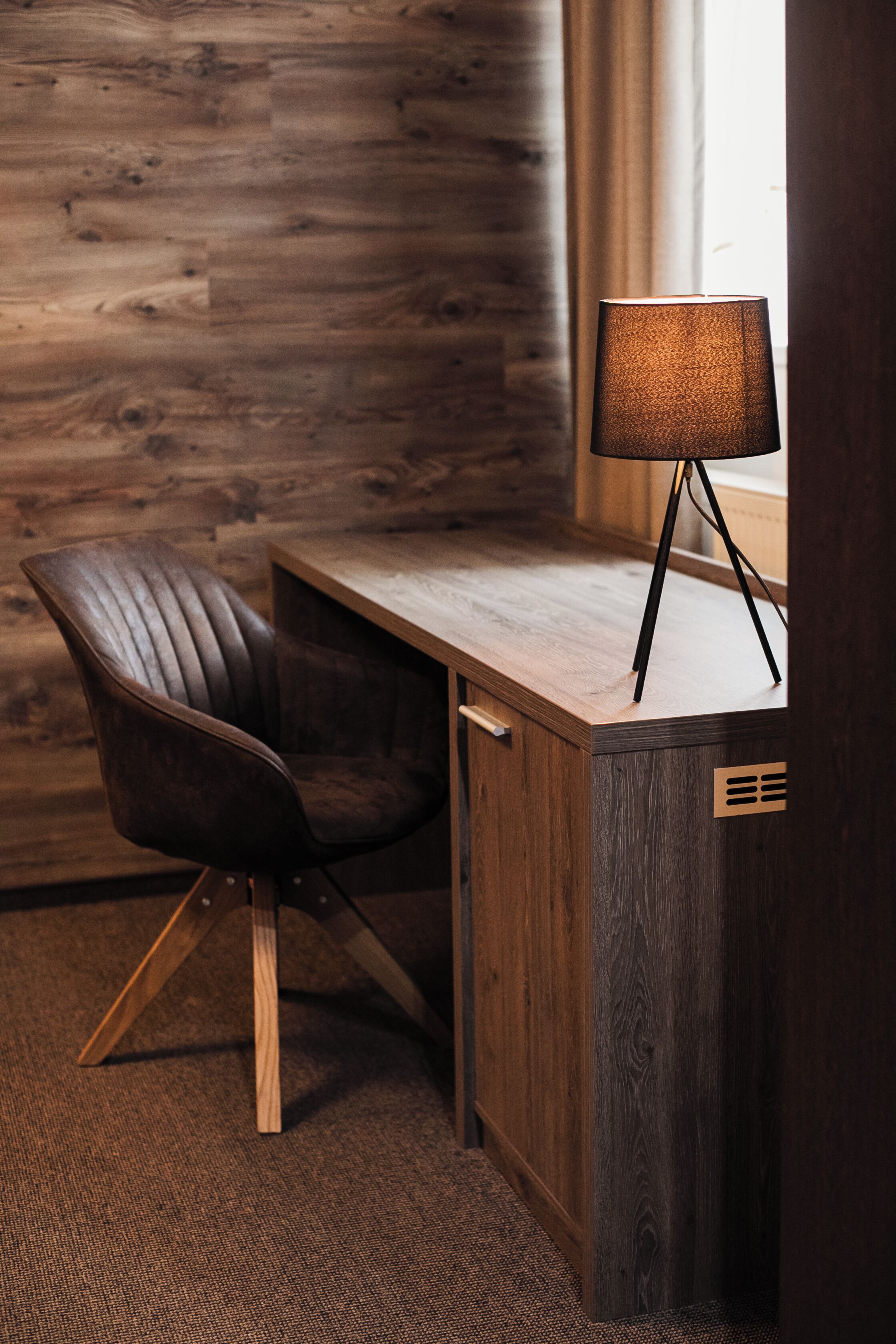 Un escritorio de madera con una lámpara, un sillón de cuero y una ventana con cortinas.