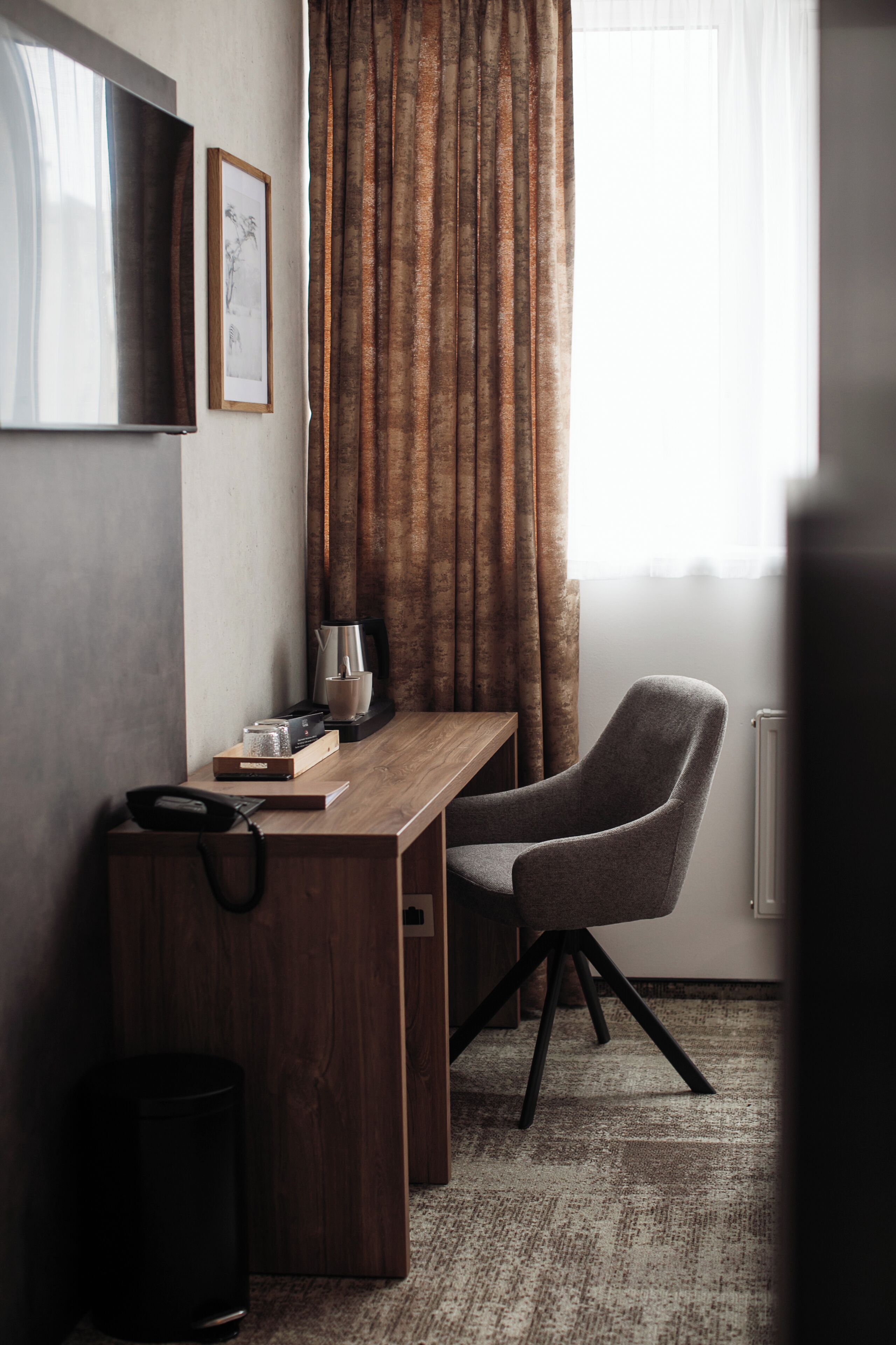 A hotel room with a wooden desk, a grey armchair, a coffee maker, and a framed picture on the wall.