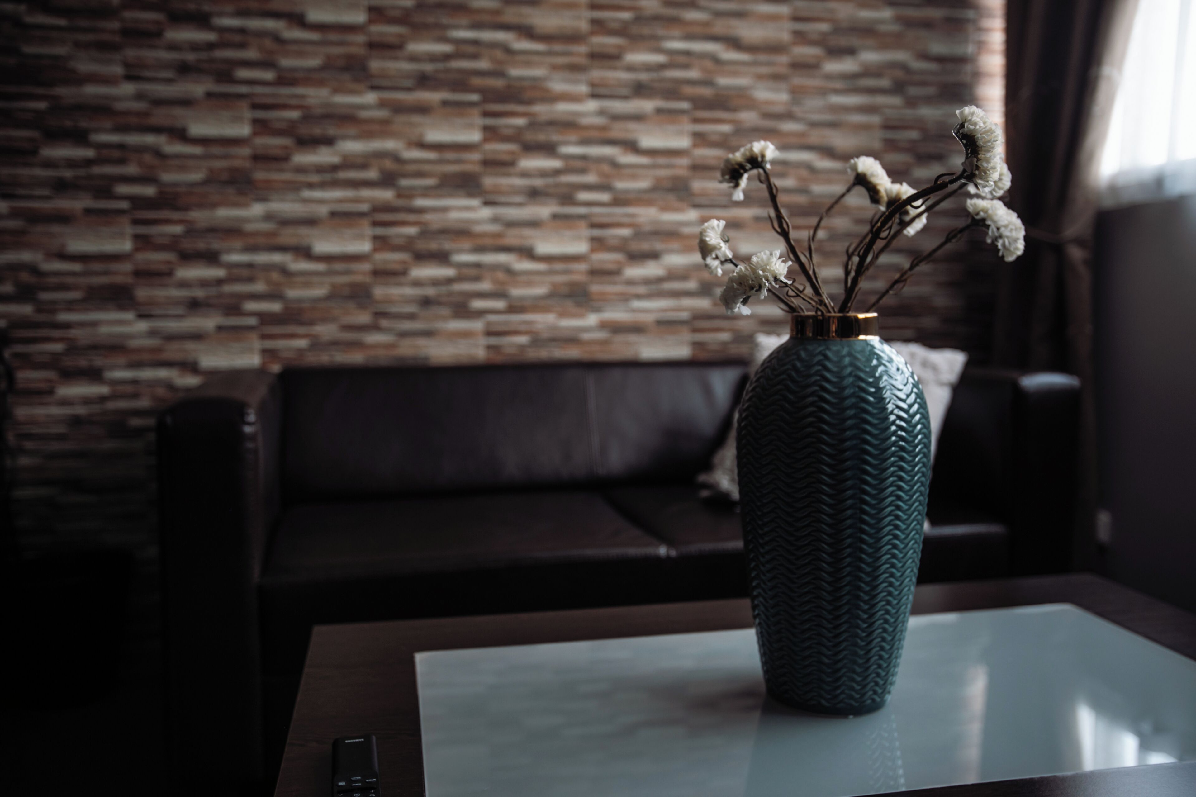 Ein modernes Wohnzimmer mit rauen Steinwänden, einem dunkelbraunen Ledersofa und einem Glastisch mit einer Vase getrockneter Blumen.