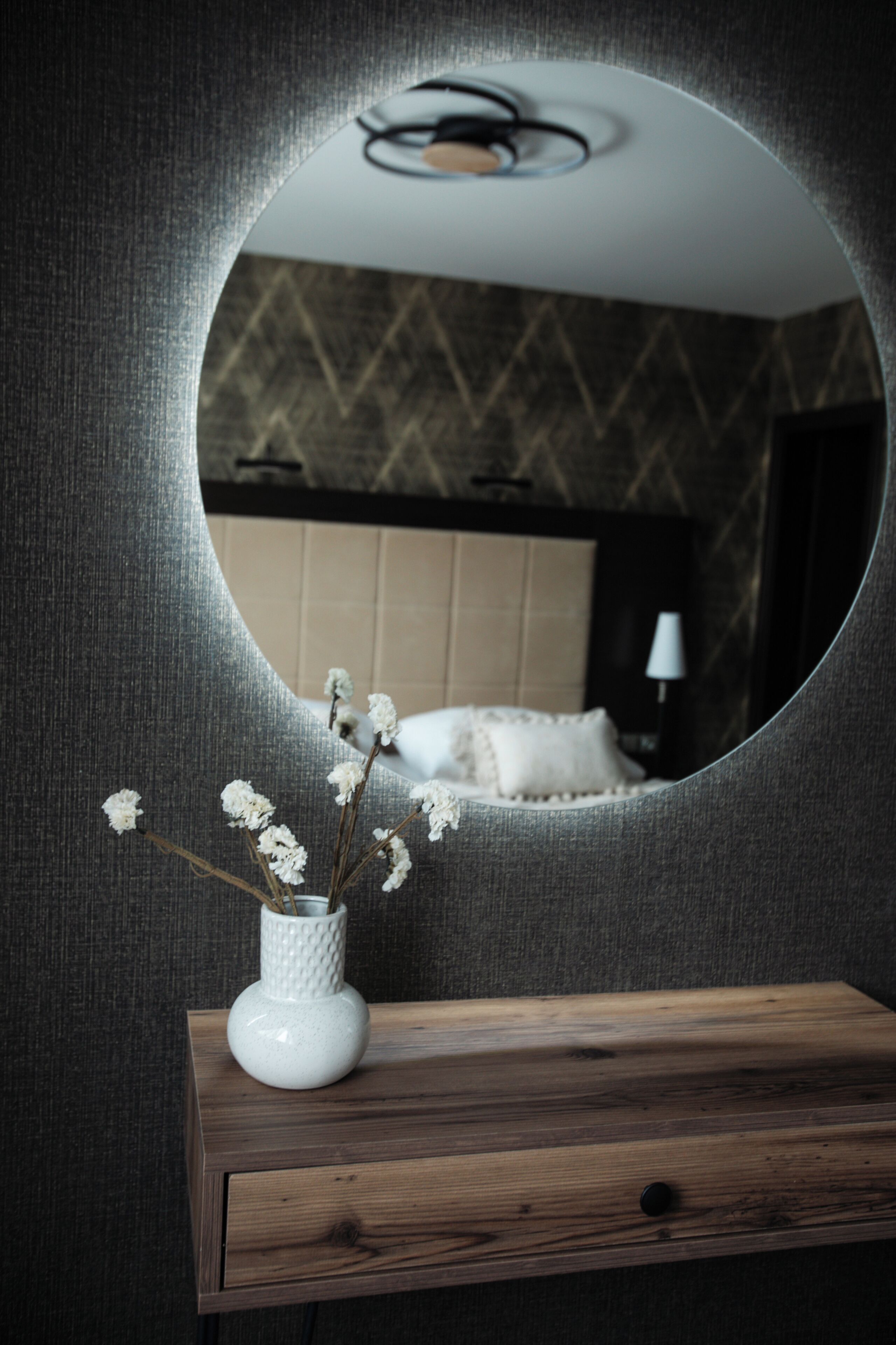 A round mirror on a wall with a wooden bedside table and a white vase with dried flowers.