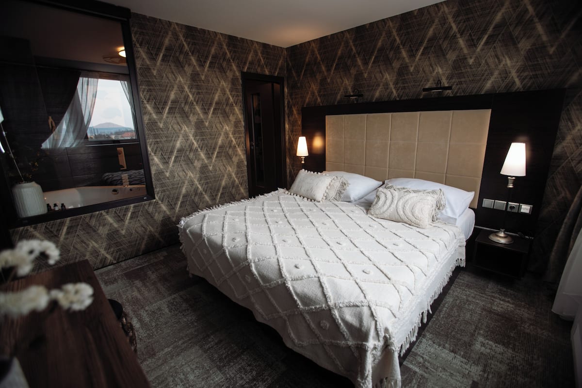 A hotel room with a large bed, a mirror, two bedside lamps, and a view of the outdoors through the window.
