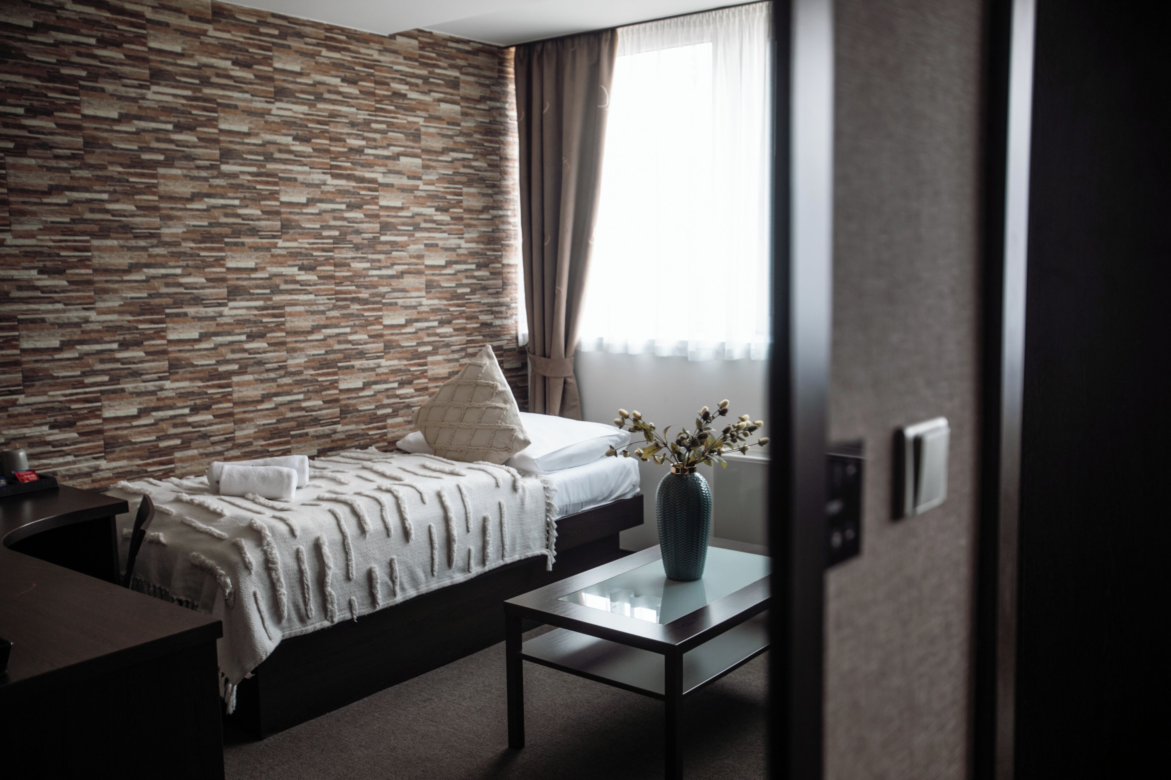 Habitación de hotel moderna con pared de diseño de piedra, cama bien tendida, una mesita con un florero y una ventana con cortinas.